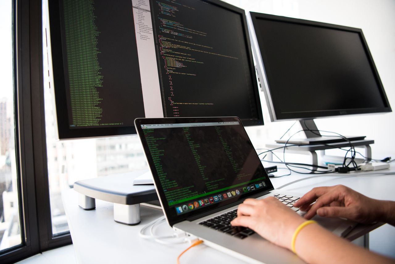 Services A developer writes code on a laptop in front of multiple monitors in an office setting.